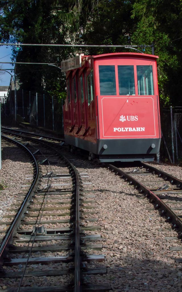"Die rote UBS Polybahn, eine Standseilbahn in Zürich, steht bereit auf den Schienen, umgeben von grüner Vegetation.