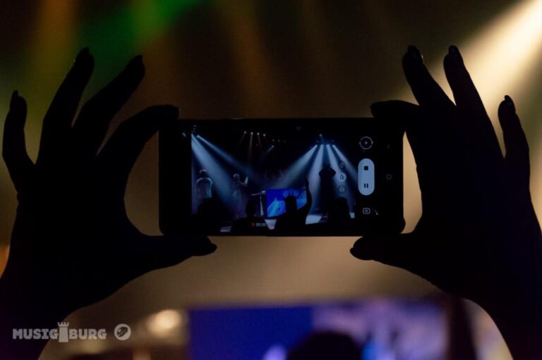 Silhouetten von Händen, die ein Smartphone halten, das ein Konzert aufnimmt, mit Bühnenlichtern, die auf dem Display des Handys zu sehen sind