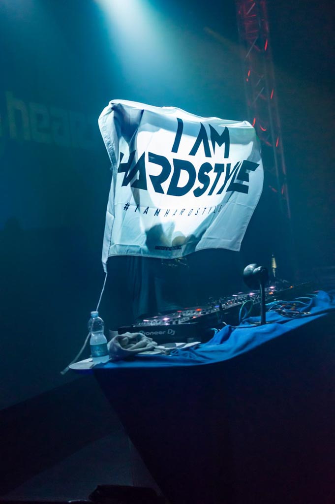 Ein Tuch mit der Aufschrift 'I AM HARDSTYLE' wird vor einem DJ-Pult in einem Club hochgehalten, mit Scheinwerfern im Hintergrund.