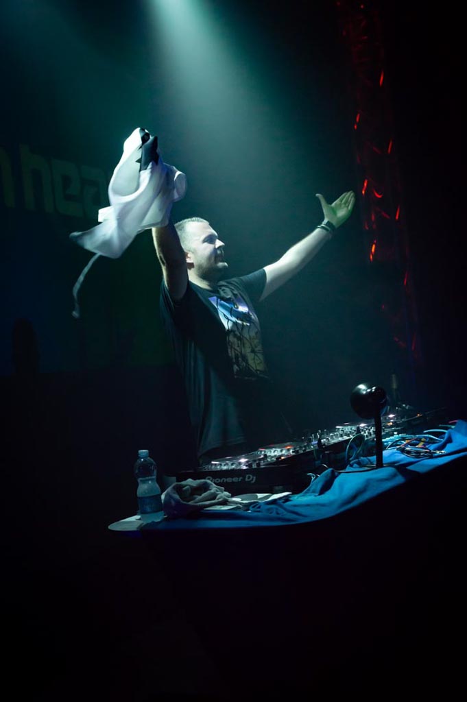Ein DJ hebt triumphierend ein Tuch mit einem Logo hoch, während er hinter einem DJ-Pult in einem Club steht.