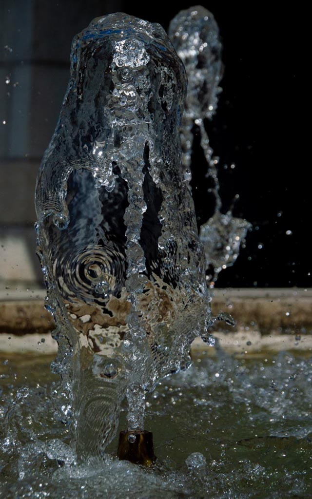 Eine Nahaufnahme einer Wassersäule, die aus einer Düse oder einem Brunnen aufsteigt. Die Wassersäule ist von zahlreichen Wasserperlen und -spritzern umgeben, die in der Luft schweben und ein dynamisches und lebendiges Bild erzeugen