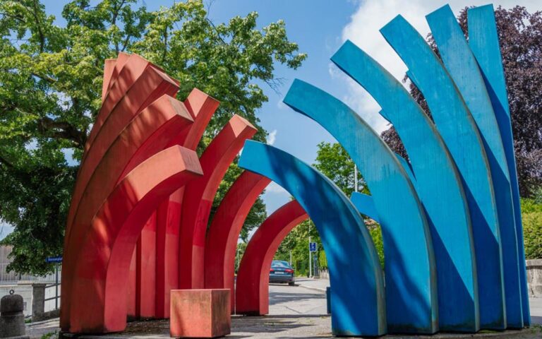 Eine moderne Skulptur im Freien mit markanten roten Bögen auf der linken Seite und einer Gruppe von blauen, vertikal ausgerichteten Elementen auf der rechten Seite. Im Hintergrund sind Bäume und ein klarer blauer Himmel zu sehen.