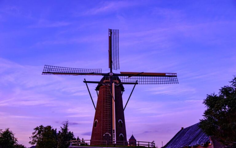 Eine traditionelle Windmühle bei Dämmerung oder kurz nach Sonnenuntergang, mit einem farbenfrohen Himmel im Hintergrund. Die Mühle ist aus roten Ziegeln gebaut und hat weisse Zierleisten an den Fenstern und Türen. Die ruhige und friedliche Stimmung wird durch die sanften Farben des Himmels und die stille Präsenz der Windmühle verstärkt.