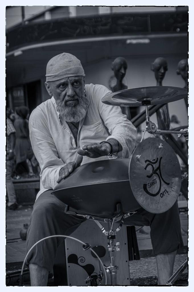 Ein älterer Strassenmusiker spielt eine Handpan mit dem Symbol "Om" darauf. Er trägt ein weisses Kopftuch und ein dunkles Oberteil und hat einen fokussierten Blick. Im Hintergrund sind unscharfe Passanten zu sehen, was darauf hindeutet, dass er auf einer belebten Strasse spielt. In Grautönen