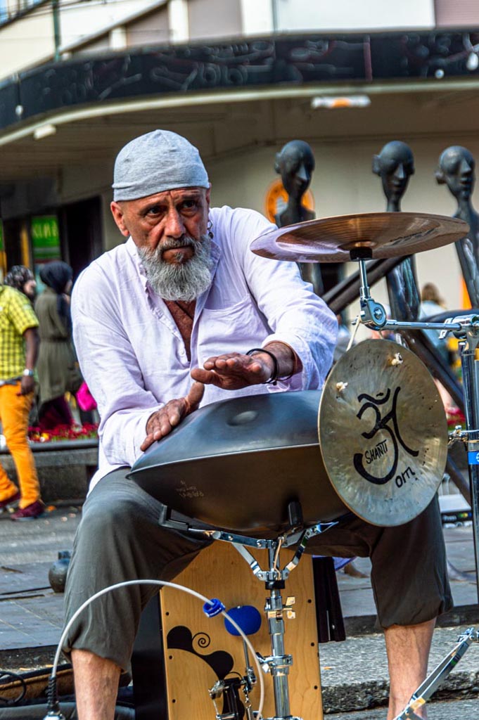 Ein älterer Strassenmusiker spielt eine Handpan mit dem Symbol "Om" darauf. Er trägt ein weisses Kopftuch und ein dunkles Oberteil und hat einen fokussierten Blick. Im Hintergrund sind unscharfe Passanten zu sehen, was darauf hindeutet, dass er auf einer belebten Strasse spielt.